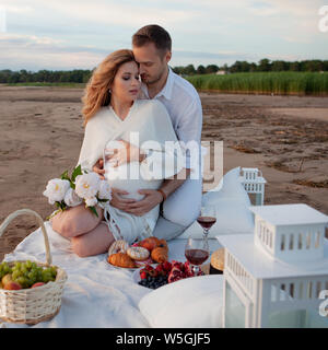 Picnic, love, tenderness. The couple sits on a plaid with pillows, flowers and fruits. A man embraces a pregnant woman from behind. Stock Photo