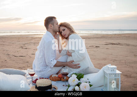 Romantic picnic at sunset. The couple is sitting on the blanket. A man gently hugs a pregnant woman. A woman looks anxiously into the distance. It see Stock Photo
