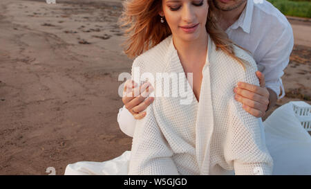 Picnic, love, tenderness. A man embraces a woman from behind by the shoulders against the background of a sandy beach. The pair is partially visible. Stock Photo