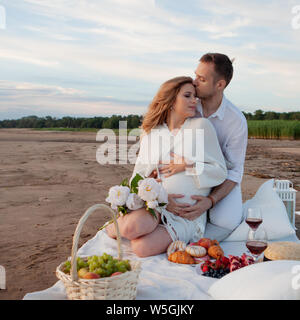 Picnic, love, tenderness. The couple sits on a plaid with pillows, flowers and fruits. A man embraces a pregnant woman from behind. Stock Photo
