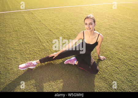 Theme sport and health. Young beautiful woman sitting doing warm-up, warming up muscles, stretching green grass. Stock Photo