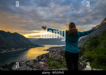 Montenegro, Beautiful young blonde woman standing with arms stretched wide above city of kotor bay in spectacular orange sunset twilight atmosphere in Stock Photo