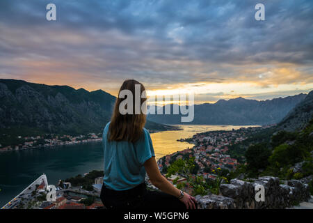 Montenegro, Young blonde beauty woman sitting on stone wall above kotor bay city in beautiful orange sunset twilight dawning atmosphere in summer vaca Stock Photo
