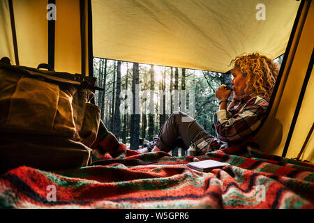 Independent beautiful curly blonde hair woman sit down outside a tent in free wild camping in the forest enjoying a cup of tea and thinking - outdoor Stock Photo