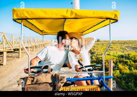 Adult people couple in love riding together a rare and funny coloured bik ein outdoor leisure activity in the nature - happy man and woman kissing eac Stock Photo