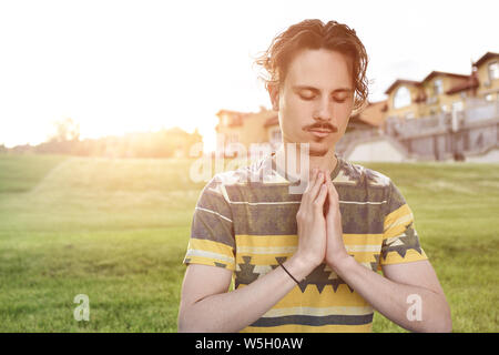 Young man meditating outdoors in the park, sitting with eyes closed and his hands together. man praying to god Stock Photo