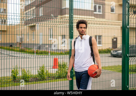 Young mixed-race basketball player with ball standing by fence of playground Stock Photo