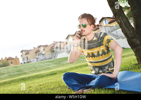 young stylish handsome guy is sitting on grass and talking on the phone resting in the park. Taking a Break Stock Photo