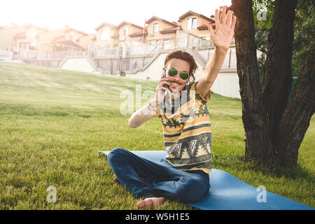 young stylish handsome man is sitting on grass, resting and talking on the phone. waves his hand welcoming friends Stock Photo