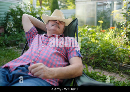 Senior hispanic man in hat sitting leaning back on chair sleeping in outdoor summer flower garden Stock Photo