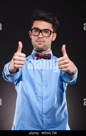 Casual guy standing and giving a double thumbs-up to the camera while wearing glasses  on gray studio background Stock Photo
