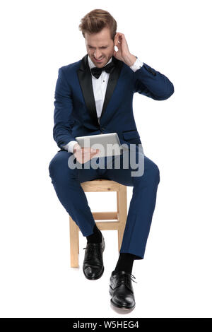 unhappy man in blue tuxedo reading bad news from notepad while sitting with hand at ear on white background Stock Photo