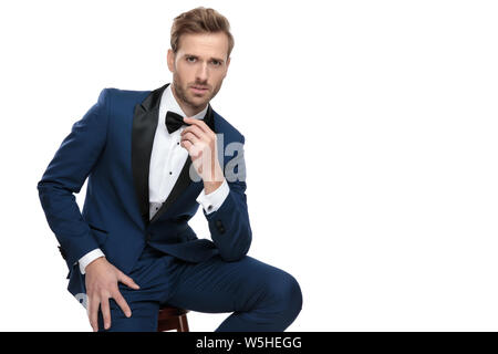 fashion man in blue suit adjusting his bowtie while sitting with hand on leg on white background Stock Photo