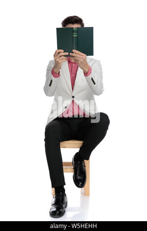 young seated elegant man reading with book covering his face, on white background Stock Photo