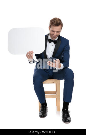 seated attractive man in blue costume texting on mobile phone while holding a blank speech bubble in his hand on white background Stock Photo