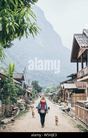 Young Caucasian blonde woman with backpack walking on the central street of Muang Ngoi village, Laos, accompanied by stray dogs. Vertical orientation Stock Photo
