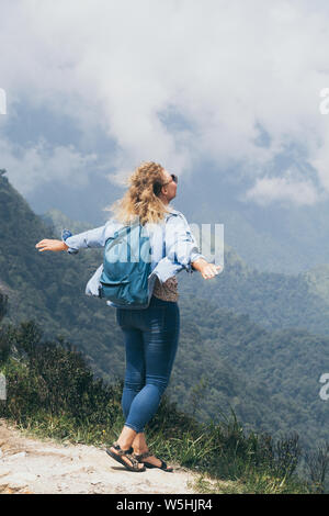 Caucasian blonde young woman drinking coffee on Tram Ton pass overlooking mountains around Sapa in Vietnam. Hands up, wind in hair. Stock Photo