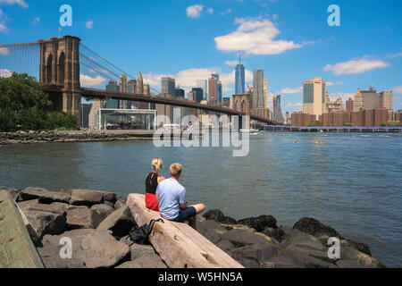 New York cityscape, view of the Brooklyn Bridge and Lower Manhattan skyline from Pebble Beach park with two young people sited in the foreground, USA. Stock Photo
