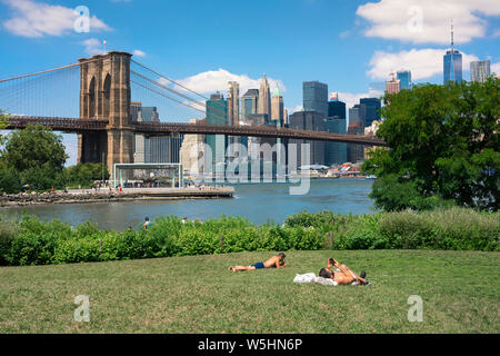 New York city summer, view of two men sunbathing in Main Street Park, Brooklyn, with the Brooklyn Bridge and Lower Manhattan skyline in the distance.