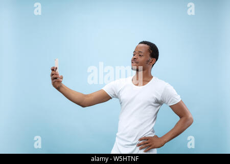 Half-length close up portrait of young african-american man in white shirt on blue background. Human emotions, facial expression, ad concept. Making selfie or content for social media, vlog. Stock Photo