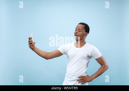 Half-length close up portrait of young african-american man in white shirt on blue background. Human emotions, facial expression, ad concept. Making selfie or content for social media, vlog. Stock Photo