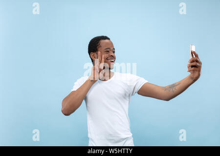Half-length close up portrait of young african-american man in white shirt on blue background. Human emotions, facial expression, ad concept. Making selfie or content for social media, vlog. Stock Photo