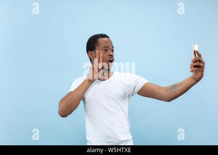 Half-length close up portrait of young african-american man in white shirt on blue background. Human emotions, facial expression, ad concept. Making selfie or content for social media, vlog. Stock Photo