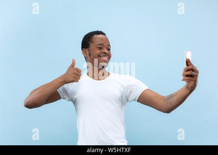 Half-length close up portrait of young african-american man in white shirt on blue background. Human emotions, facial expression, ad concept. Making selfie or content for social media, vlog. Stock Photo