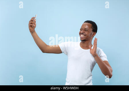 Half-length close up portrait of young african-american man in white shirt on blue background. Human emotions, facial expression, ad concept. Making selfie or content for social media, vlog. Stock Photo