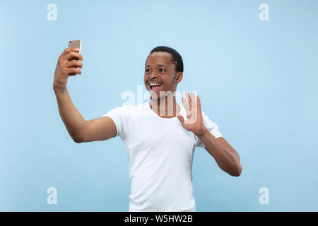 Half-length close up portrait of young african-american man in white shirt on blue background. Human emotions, facial expression, ad concept. Making selfie or content for social media, vlog. Stock Photo