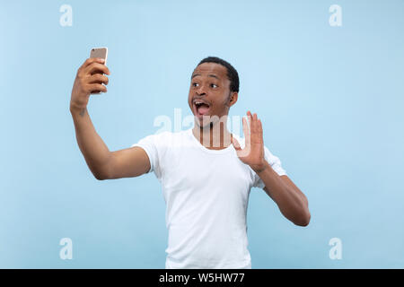 Half-length close up portrait of young african-american man in white shirt on blue background. Human emotions, facial expression, ad concept. Making selfie or content for social media, vlog. Stock Photo