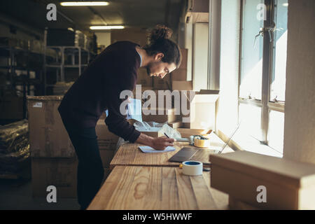 Man making note of the order he is packing to delivery. Young man preparing parcel box of product for delivery to customer. Stock Photo