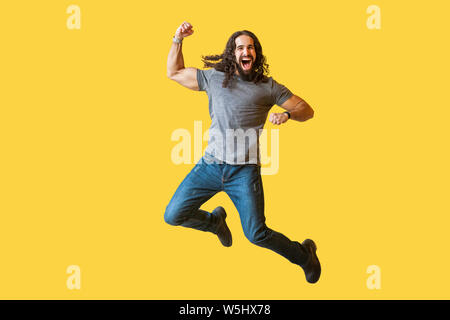 Portrait of happy rejoicing bearded young man with long curly hair in casual grey tshirt jumping and celebrating his vivtory with amazed excited face. Stock Photo