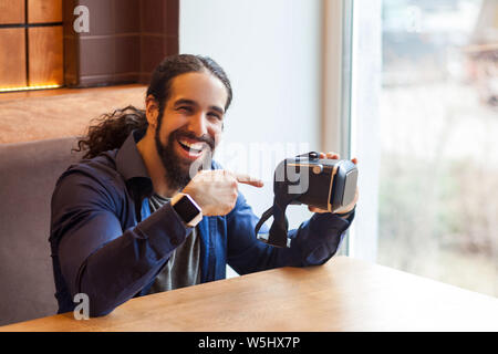 Portrait of happy bearded young adult man in casual style sitting in cafe, holding vr and pointing finger on simulator with happy face, looking at cam Stock Photo