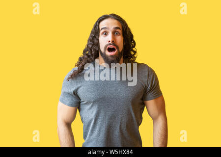 Portrait of shocked bearded young man with long curly hair in grey tshirt standing and looking at camera with open mouth and big eyes and ubelievable Stock Photo