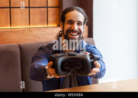 Portrait of happy bearded young adult man in casual style sitting in cafe, holding and showing vr back view, looking at camera and toothy smile. Indoo Stock Photo