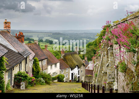 Stone houses along the steep cobbled street of Gold Hill in Shaftsbury England with view of farm fields in rain Stock Photo