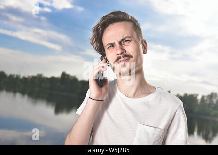Portrait of angry confused aggressive in bad mood guy irritably speaks on the phone on the background of the lake Stock Photo