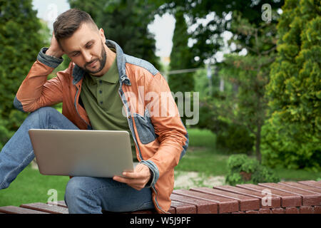 Young, successful freelancer working on laptop, sitting on wooden bench in spring green garden. Handsome, concentrated man in stylish clothing looking at laptop. Stock Photo