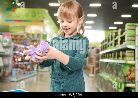 Modern toys for development in store of children toys. Happy pretty girl playing with violet kinetic sand in shopping centre. Child standing in playing room of store. Stock Photo