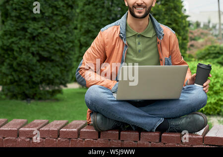 Unrecognizable young man in green t shirt, jeans and leather jacket with laptop and paper cup of coffee. Cheerful freelancer sitting with crossed legs on wooden bench. Stock Photo