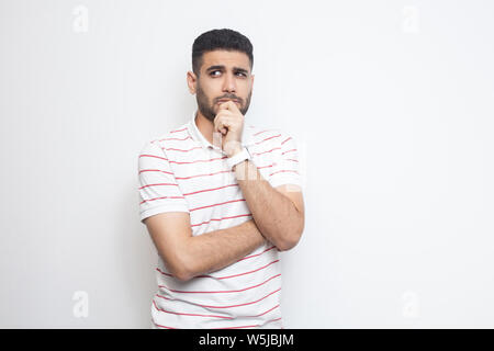 Portrait of thoughtful handsome bearded young man in striped t-shirt standing, looking away and thinking what to do. indoor studio shot, isolated on w Stock Photo