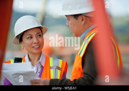Two engineers in high visibility vests and hardhats discuss the plans for a new building. Stock Photo
