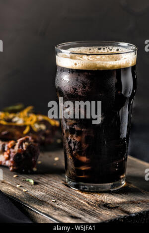 Dark beer in a steamed glass and grilled pork ribs in tomato sauce with chili pepper on a dark wooden Board. The food of the restaurant. Side view. Stock Photo