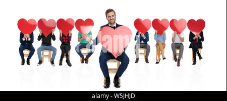 happy elegant man in tuxedo and bowtie welcomes the love while sitting in fron of a large team of in love people Stock Photo