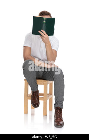 casual man sitting and reading a book covering his face with it on white background Stock Photo