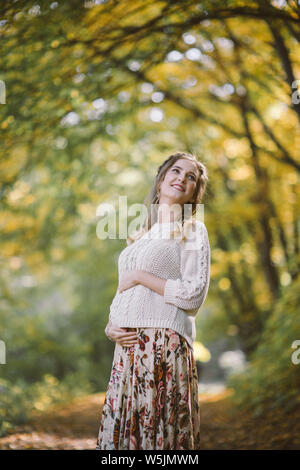 Smiling pregnant woman in sweater standing in autumn park Stock Photo