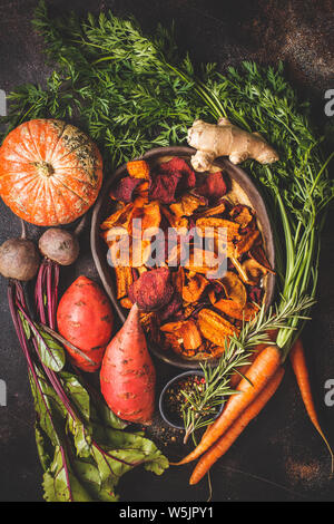 Dish of healthy vegetable chips from beets, sweet potatoes and carrots with ingredients on dark background, top view. Stock Photo