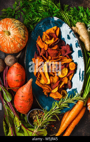Dish of healthy vegetable chips from beets, sweet potatoes and carrots with ingredients on dark background, top view. Stock Photo