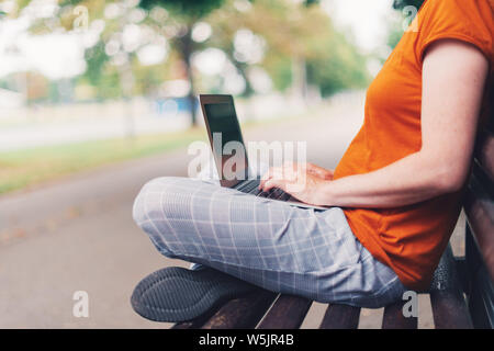Woman using laptop computer on street bench, mock up screen. Adult caucasian female blogger is typing on keyboard, working anywhere away from home off Stock Photo